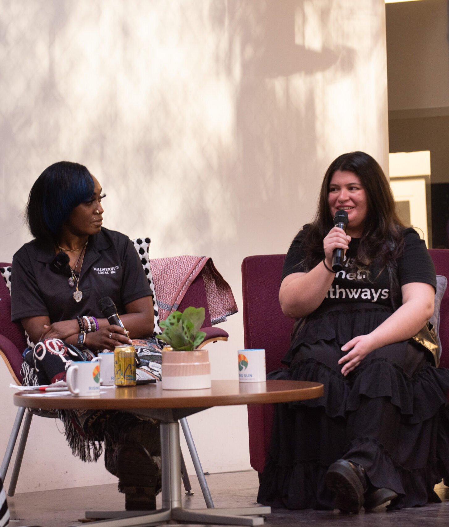 Two women seated in armchairs participate in a panel discussion at a Rising Sun Center for Opportunity event. One panelist holds a microphone and speaks while the other panelist, seated to the left, looks at them.