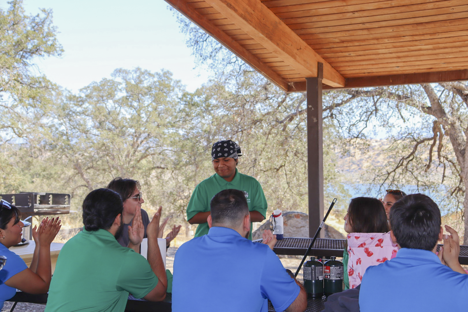 Neftaly Cervantes stands and speaks to a group of Climate Careers peers seated at a picnic table under a wooden pavilion, with trees and hills in the background.