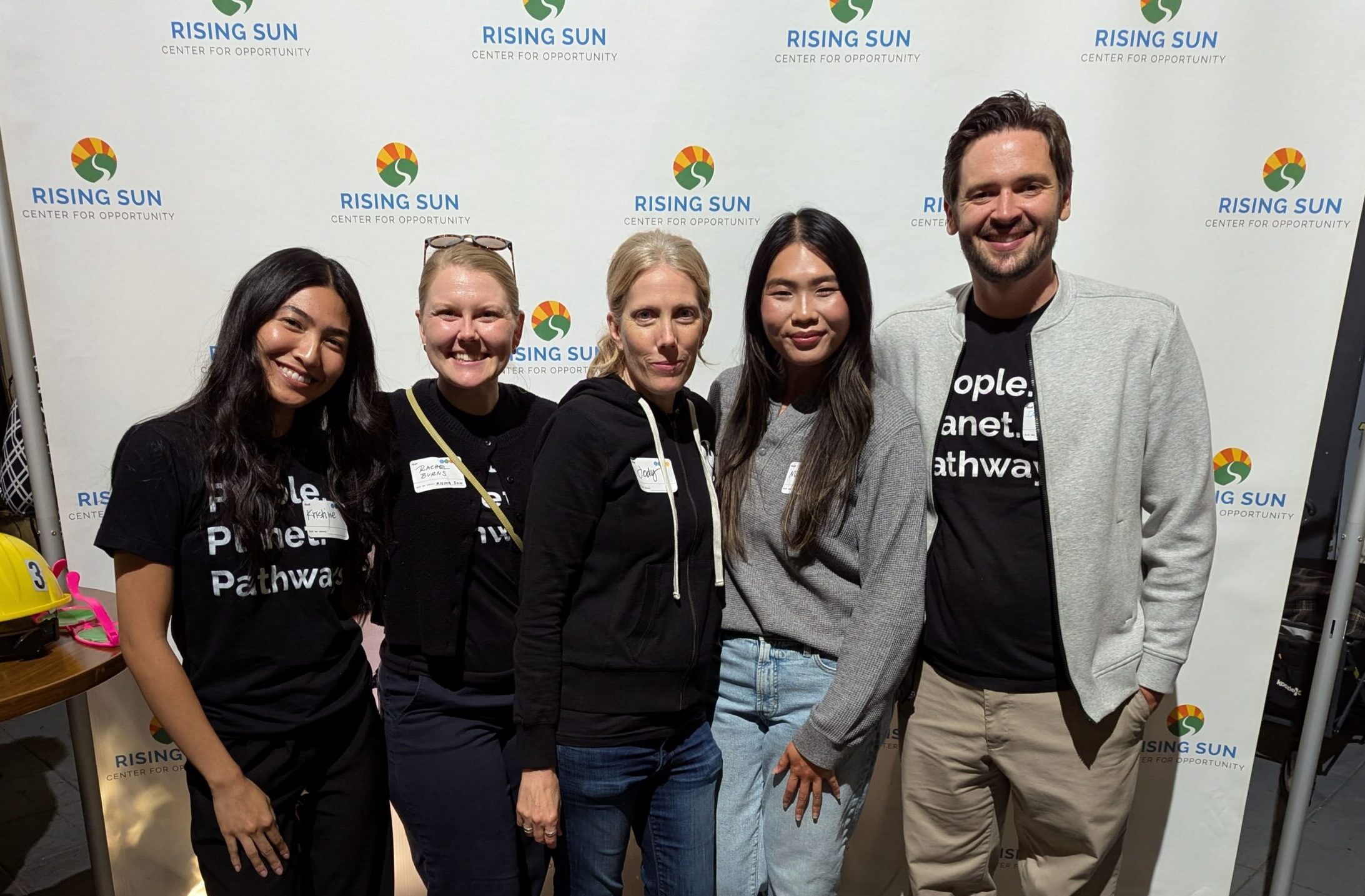 Five people smile together in front of a Rising Sun Center for Opportunity step-and-repeat banner. Three are wearing black 